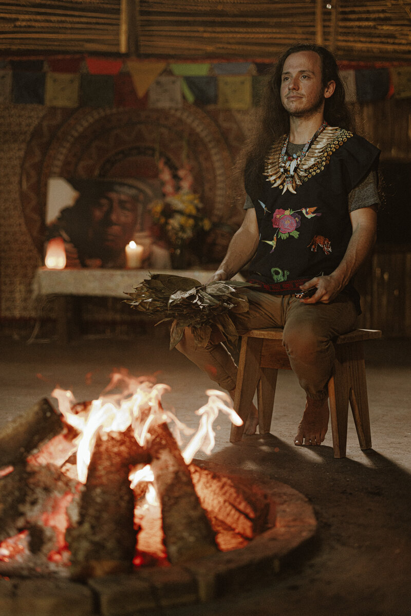 Michael seated with ceremonial leaves beside a fire in the maloka